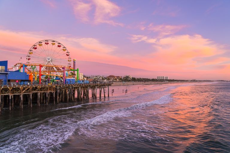 Santa Monica Pier featuring Pacific Park's Ferris wheel and roller coaster over the ocean during a pink and orange sunset.